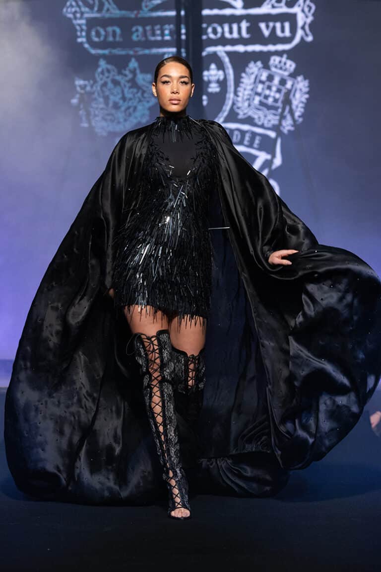 A model walks the On Aura Tout Vu couture runway wearing a short black dress with sparkling fringe, paired with a voluminous black organza cape coat and lace-up thigh-high boots