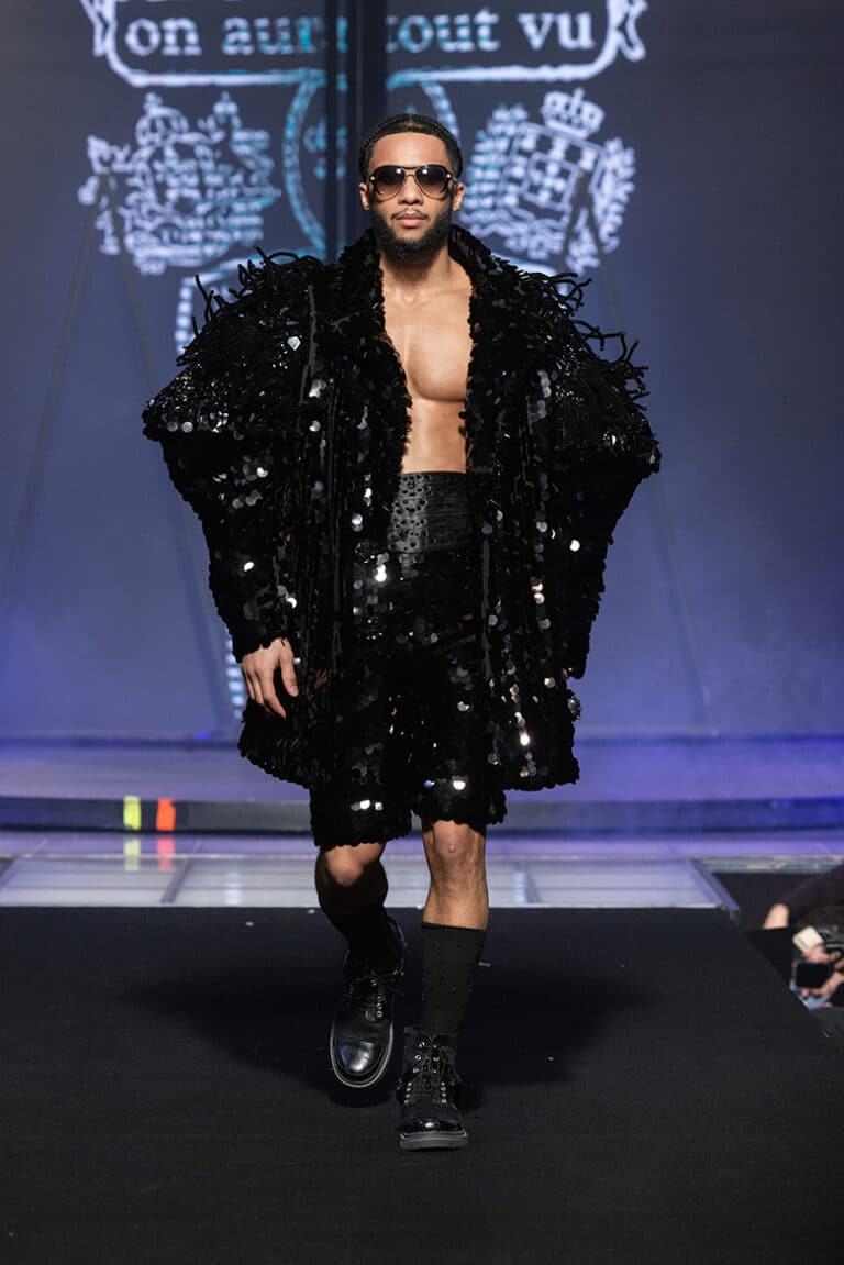 A male model walk on aura tout vu show wearing black sequinned shorts, satin belt, and a long coat embroidered with sequins and feathers