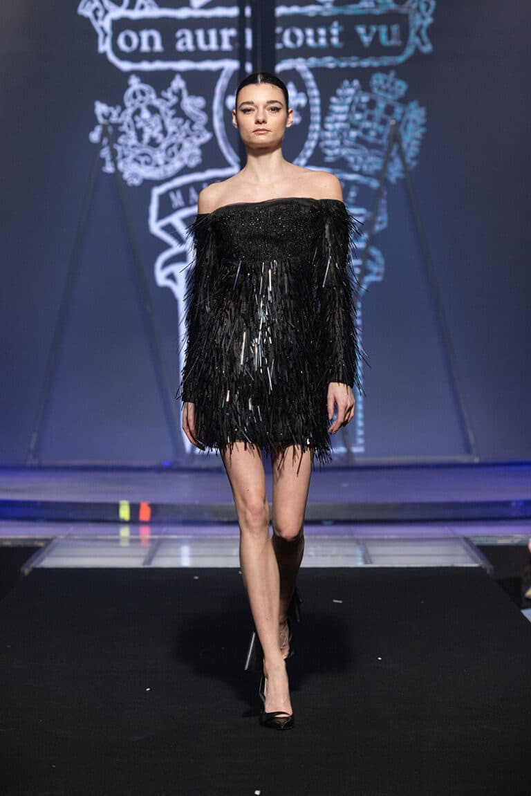 A model walks the On Aura Tout Vu couture runway wearing a short black dress with sparkling fringe, paired with lace-up thigh-high boots