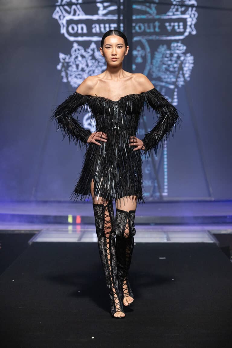 A model walks the On Aura Tout Vu couture runway wearing a short black dress with sparkling fringe, paired with lace-up thigh-high boots