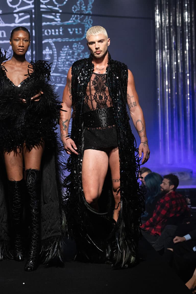 A male model walk on aura tout vu show wearing black sequinned micro-shorts, a transparent lace top with baroque patterns, and a long sleeveless coat decorated with sequins and feathers