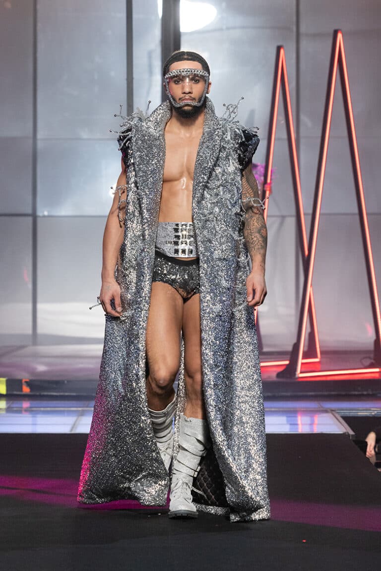 A male model walk on aura tout vu show wearing silver sequinned micro-shorts, and sleeveless coat with sequins and spikes.