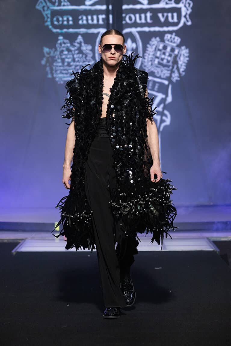 A male model walk on aura tout vu show wearing black pants and black satin belt, long sleeveless coat embroidered with sequins and feathers