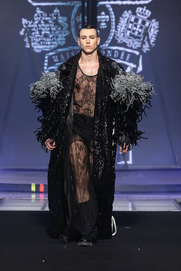 A male model walk on aura tout vu show wearing transparent black lace top and pants with baroque patterns, and a long coat decorated with sequins and feathers